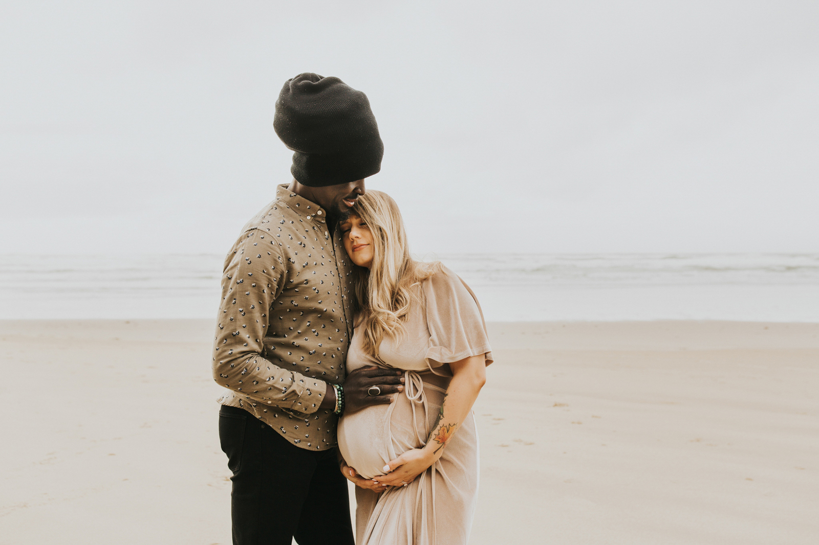 Interracial couple snuggle with dad's hand on mom's belly during their maternity session on the oregon coast with Meg Ross Photography