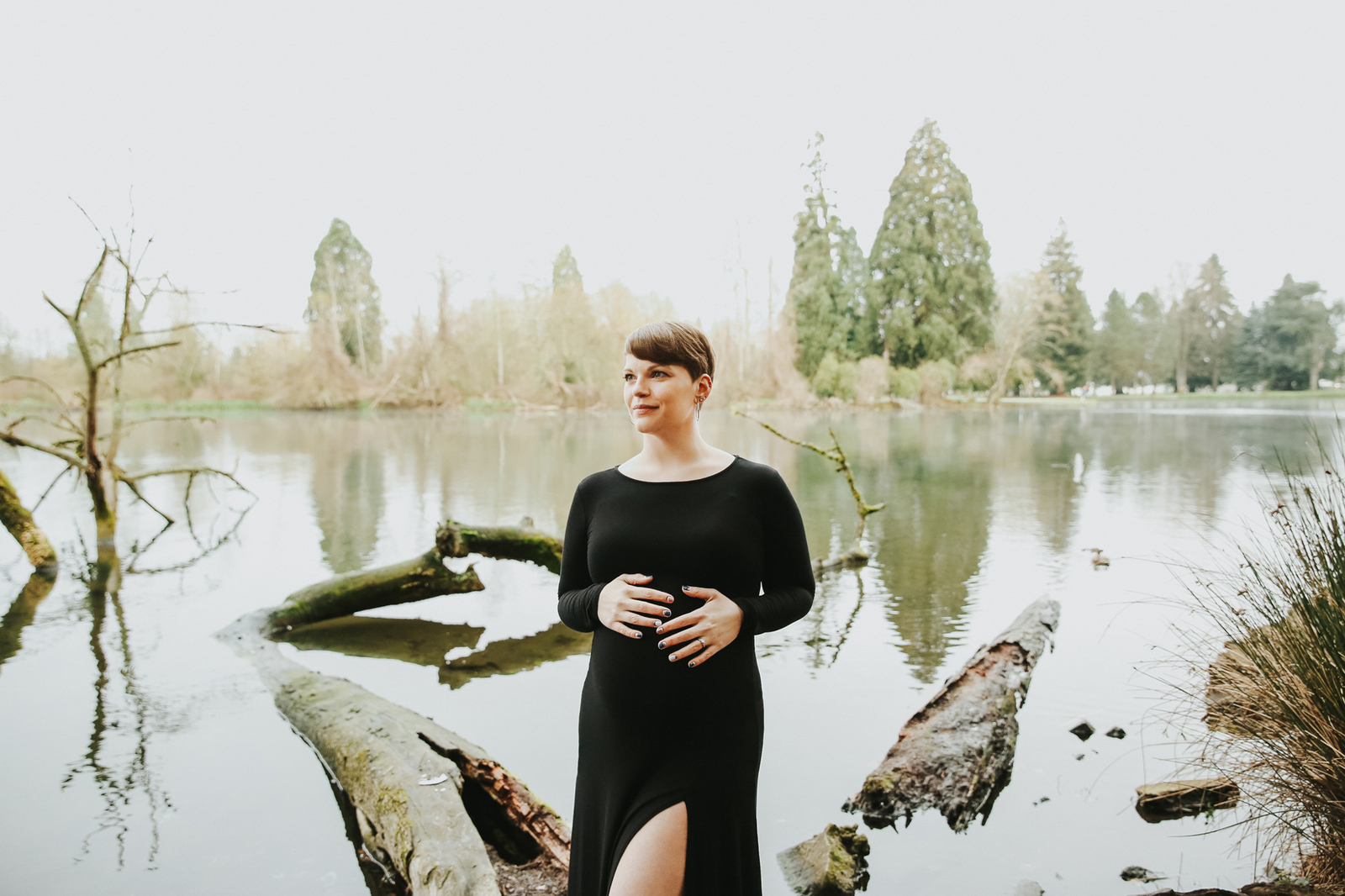 Pregnant mama in a black dress holds her bump and looks off into the distance with trees and water in the background during into the distance at Crystal Rhododendron Garden with Portland Maternity Photographer.