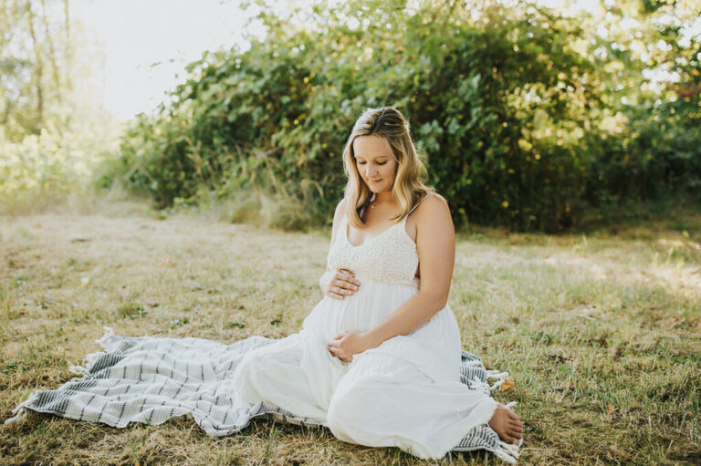 Pregnant Nicole is sitting down on a blanket, looking down, and holding her bump in a white dress during her maternity session in Yamhill, Oregon