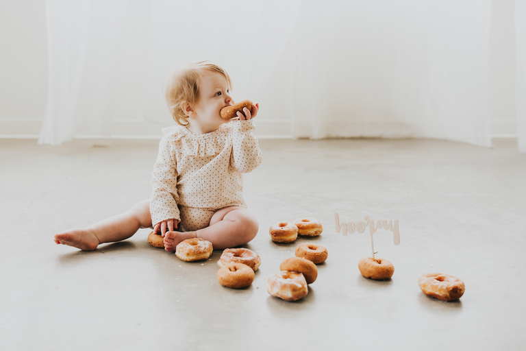 A redheaded tot sits on the floor with a donut in her mouth and donuts on the floor around her during her Portland Oregon first birthday donut smash.