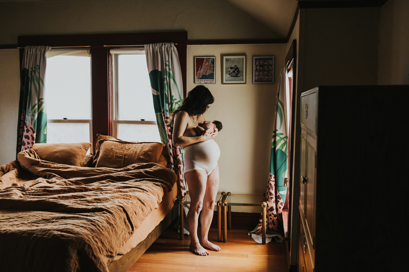 Postpartum mom in mesh underwear nurses baby, standing in a window, in the middle of her bedroom in Portland Oregon